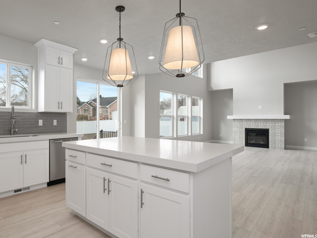 kitchen featuring a fireplace, natural light, stainless steel dishwasher, pendant lighting, white cabinetry, light countertops, and light hardwood floors