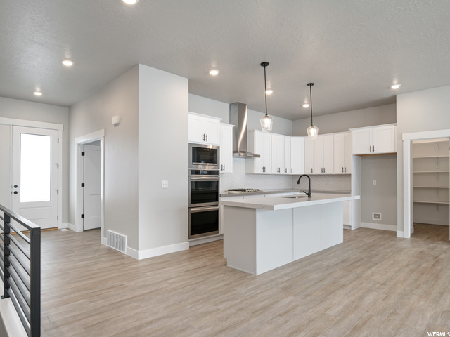kitchen with a wealth of natural light, double oven, range hood, pendant lighting, light countertops, kitchen island sink, light hardwood floors, and white cabinetry