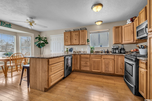 kitchen with a ceiling fan, a breakfast bar area, natural light, stainless steel dishwasher, microwave, gas range oven, dark countertops, brown cabinetry, and light hardwood floors