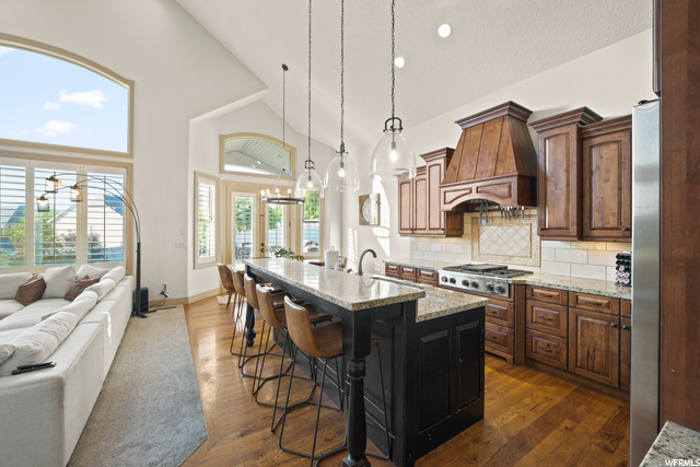 Kitchen with natural light, a kitchen breakfast bar, gas stovetop, stainless steel finishes, fume extractor, light granite-like countertops, an island with sink, light hardwood flooring, and pendant lighting
