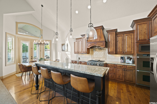 Kitchen featuring a breakfast bar, hardwood floors, natural light, gas stovetop, double oven, exhaust hood, stone countertops, a center island with sink, and pendant lighting