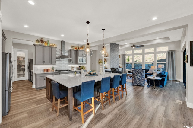 Kitchen with light hardwood flooring, decorative light fixtures, backsplash, ceiling fan, a center island, appliances with stainless steel finishes, a tray ceiling, kitchen island with sink, and wall chimney exhaust hood