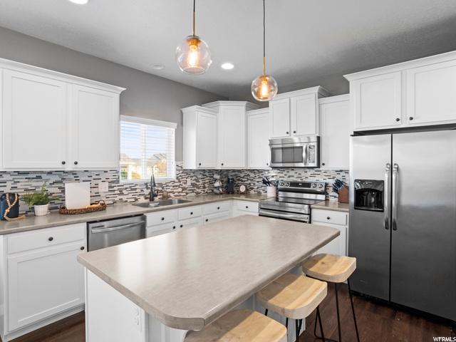 Kitchen featuring a kitchen island, stainless steel appliances, white cabinets, dark hardwood floors, light countertops, hanging light fixtures, and backsplash