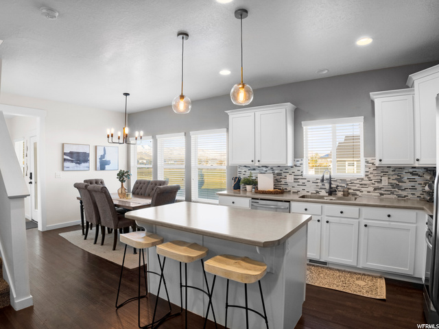 Kitchen featuring stainless steel dishwasher, hanging light fixtures, backsplash, white cabinets, light countertops, a center island, and dark hardwood flooring