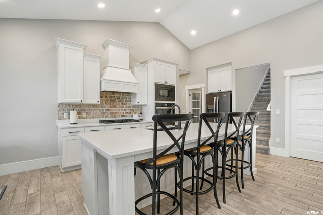Kitchen featuring backsplash, white cabinets, light countertops, premium range hood, a center island, light hardwood flooring, lofted ceiling, stainless steel appliances, and a high ceiling