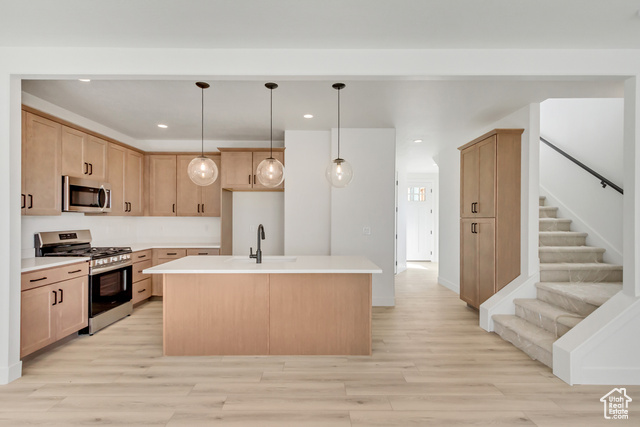 Kitchen featuring light brown cabinetry, decorative light fixtures, appliances with stainless steel finishes, light hardwood / wood-style floors, and a center island with sink