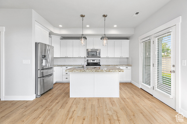 Kitchen featuring light wood-type flooring, an island with sink, hanging light fixtures, appliances with stainless steel finishes, and white cabinets