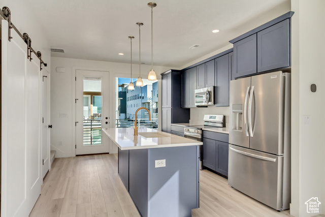 Kitchen with appliances with stainless steel finishes, a barn door, light hardwood / wood-style floors, pendant lighting, and a kitchen island with sink