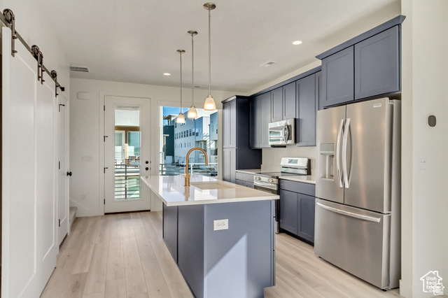 Kitchen featuring a kitchen island with sink, light hardwood / wood-style flooring, hanging light fixtures, stainless steel appliances, and a barn door