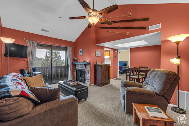 Carpeted living room featuring a skylight, ceiling fan, a stone fireplace, and high vaulted ceiling