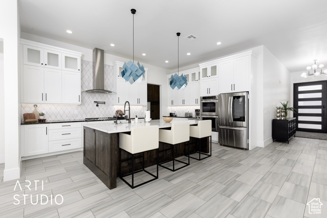 Kitchen featuring stainless steel appliances, wall chimney range hood, decorative light fixtures, a center island with sink, and white cabinets