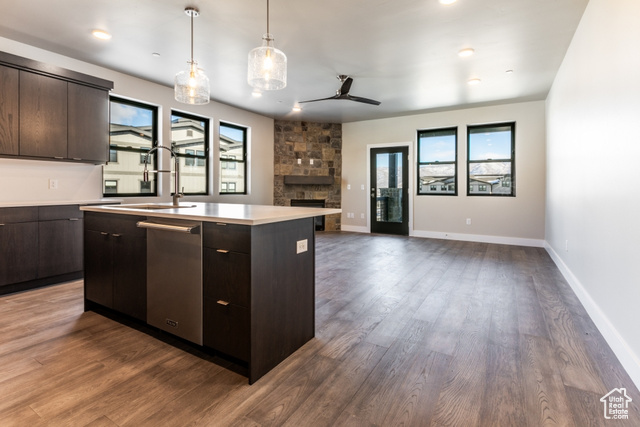 Kitchen featuring dark hardwood / wood-style flooring, dark brown cabinetry, a kitchen island with sink, ceiling fan, and a stone fireplace